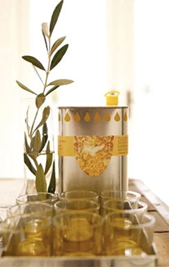 A tin of olive oil with cups of olive oil in the foreground and a stem from an olive tree in the background